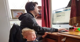 A man sits at a computer while his toddler baby cries at his side.