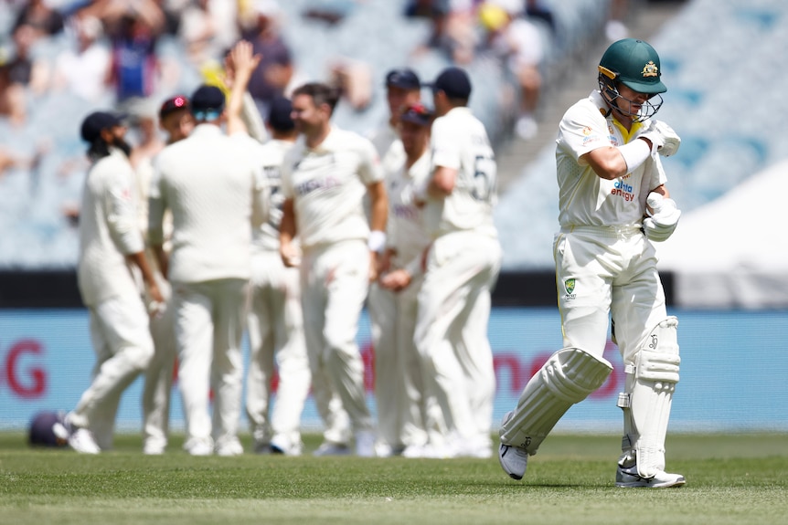 Australia batter Marcus Harris tucks his bat under his arm as he leaves the field. England players gather in the background.