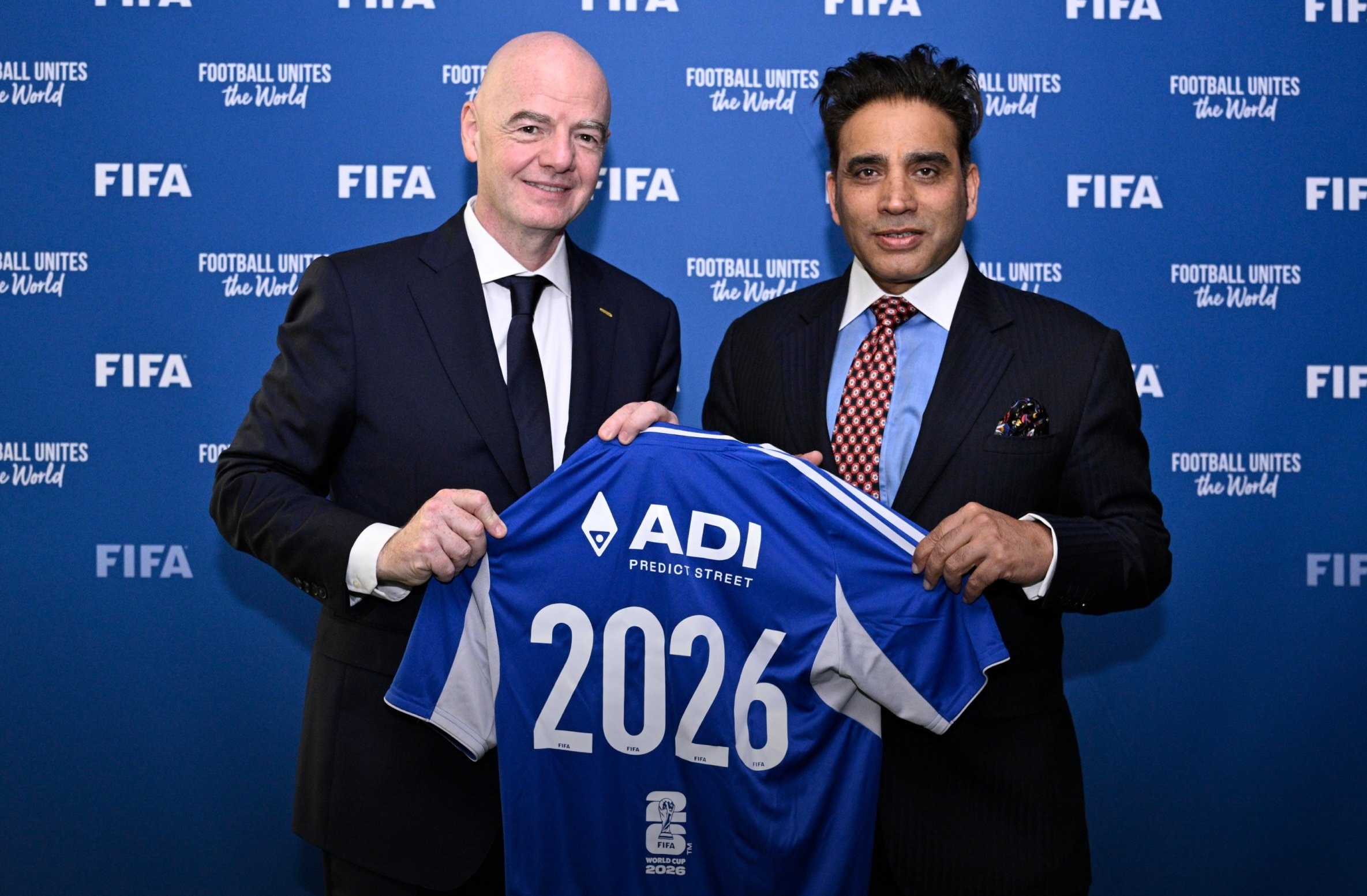 Two men in suits, at a press conference, hold up a football jersey with ADI Predict Street and the number "2026" on the back.