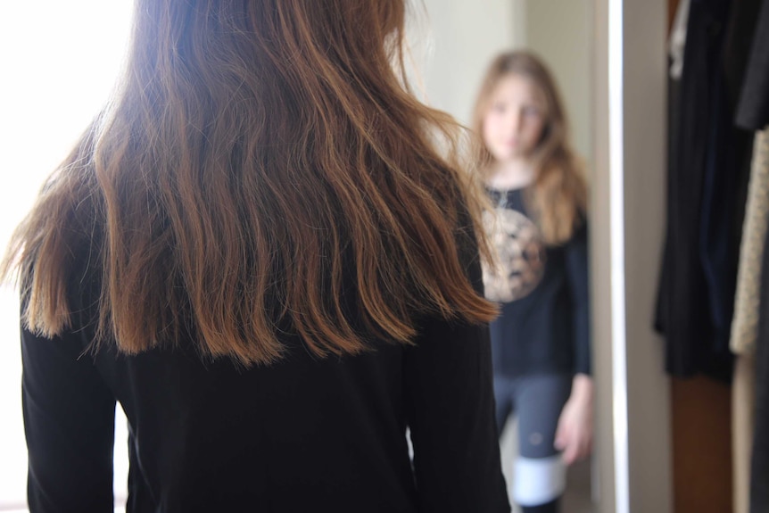 A young girl with her back to camera looks into her reflection in a mirror.