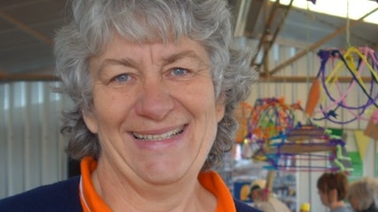 A woman stands in front of a local craft stall wearing an orange t-shirt, navy cardigan and name badge.