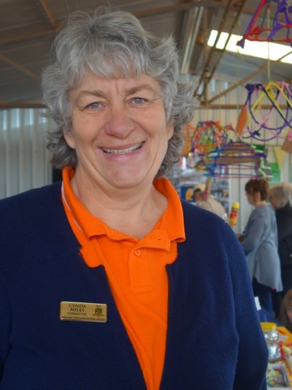 A woman stands in front of a local craft stall wearing an orange t-shirt, navy cardigan and name badge.