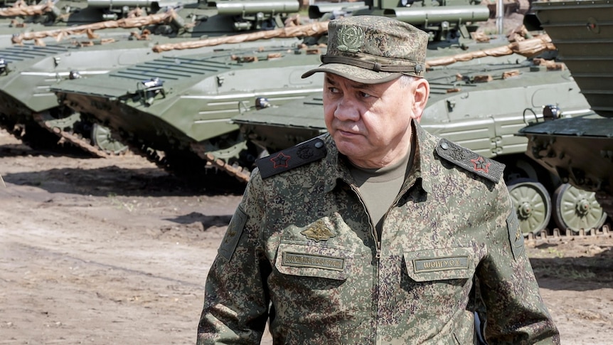 Sergei Shoigu, a round-faced older man in military uniform and cap, walks past a row of tanks