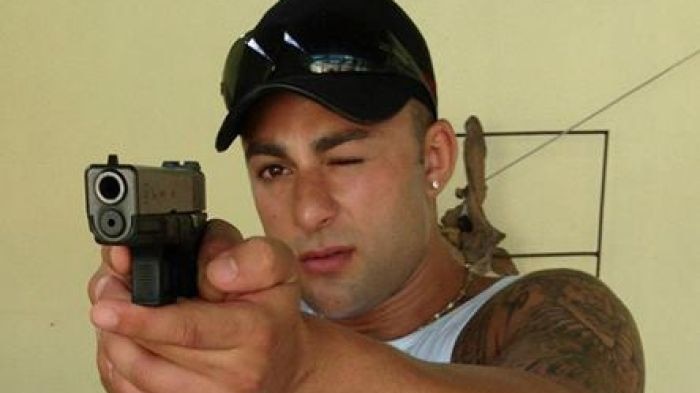 A man points a gun directly at the camera. He is wearing a cap with sunglasses on top, and has one eye closed.