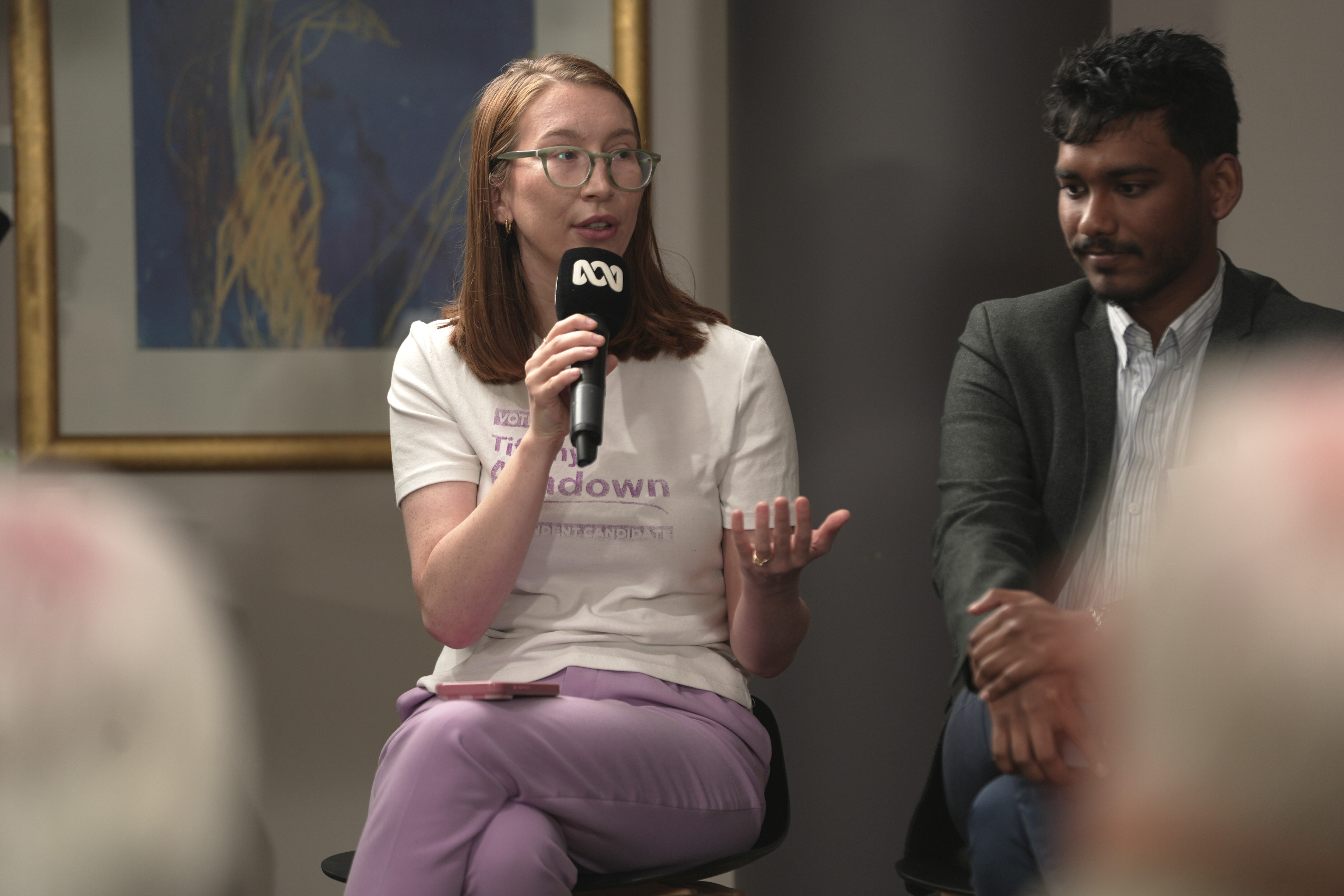 A woman sits on a chair holding an ABC microphone while speaking to an audience