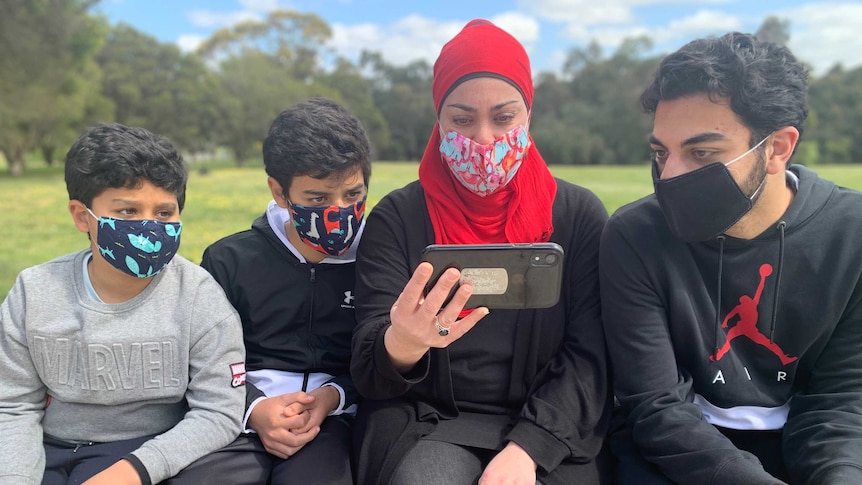 A family of two young boys, a woman and an older teenager, all wearing face masks and looking at a phone.