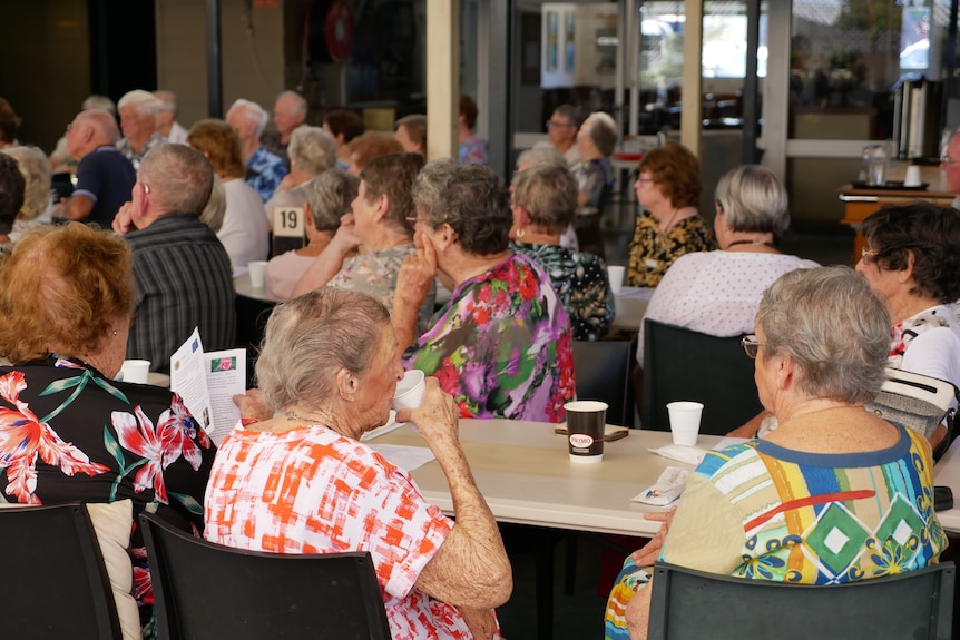 Seniors gather around tables in a well lit function room looking towards a speaker.