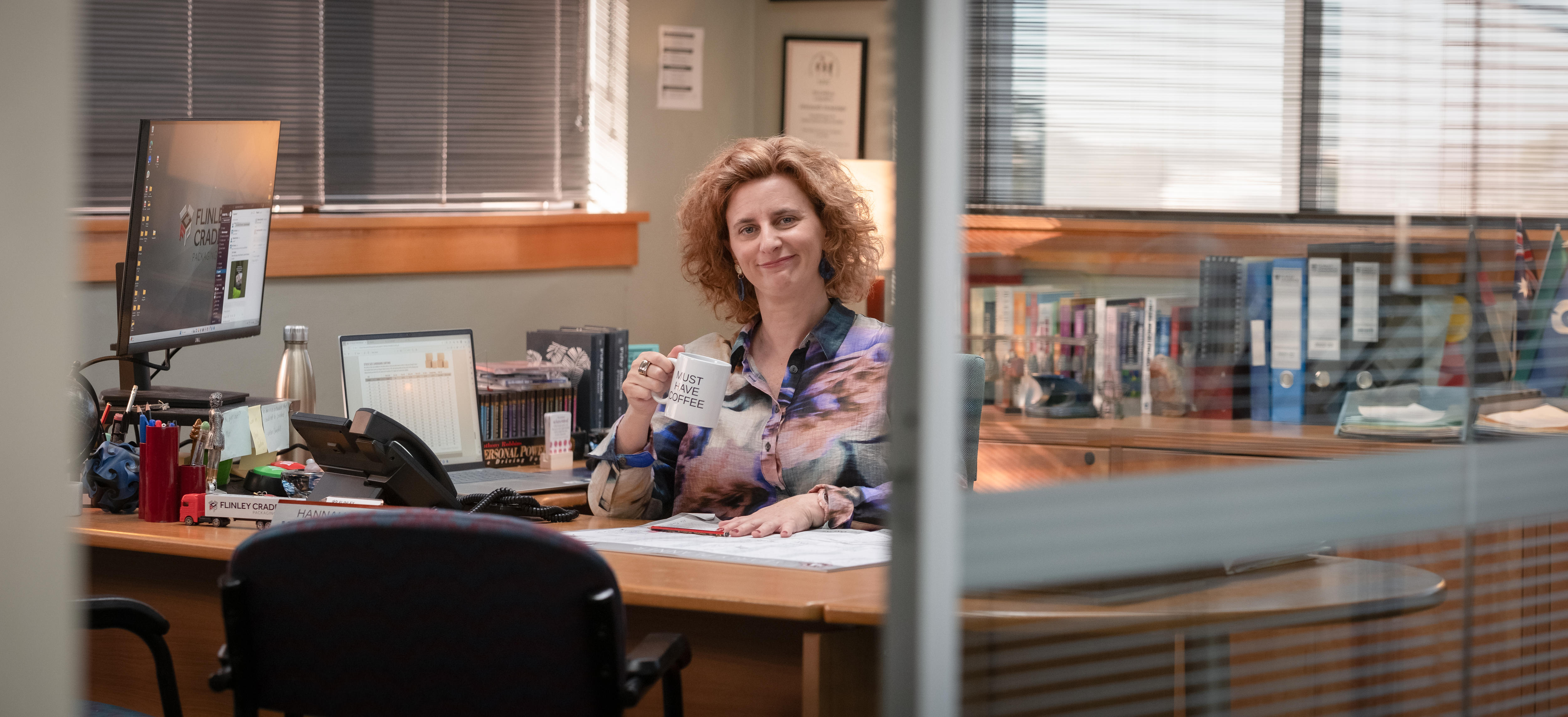 A woman sits behind a desk holding a coffee mug and smiles at the camera
