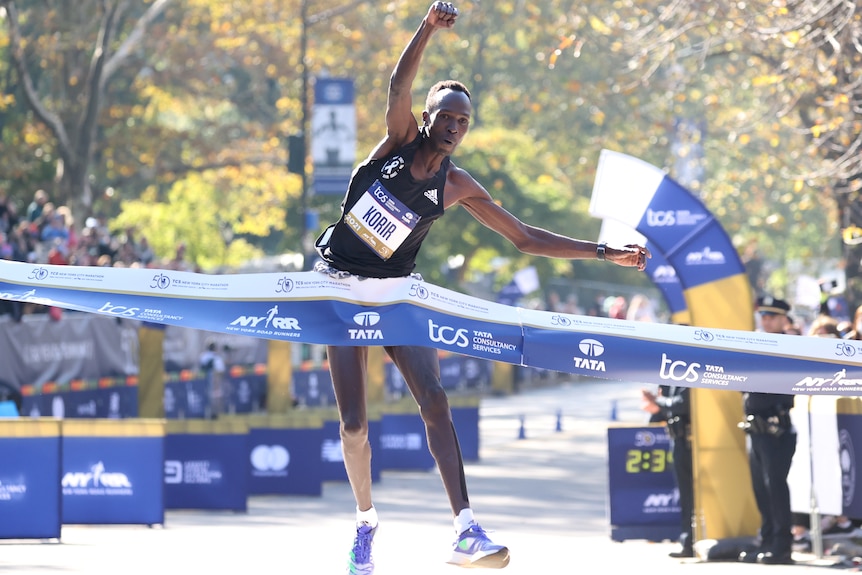 Albert Korir jumps with one hand up in the air as he passes through a blue finishing banner