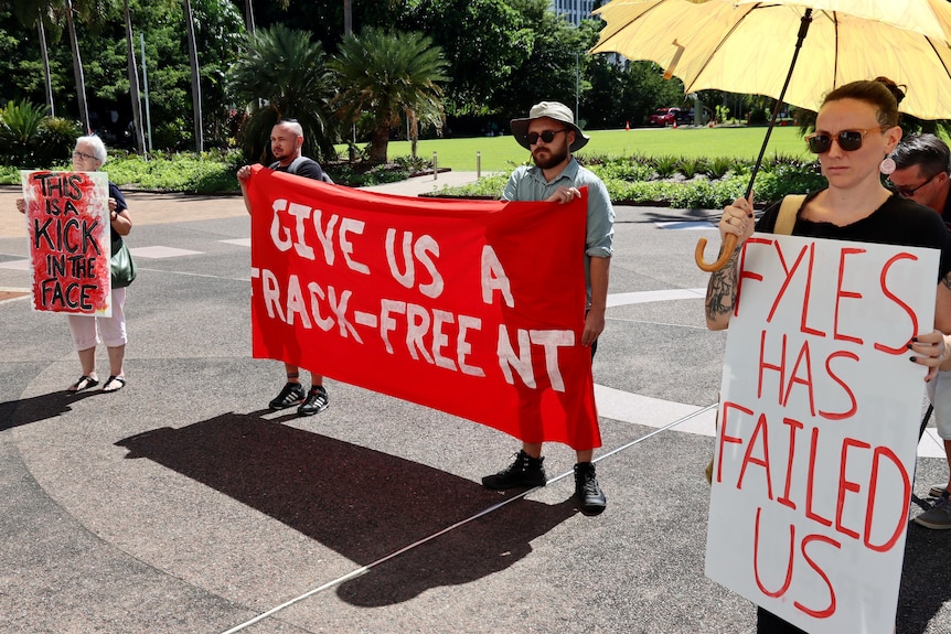 Four people stand on a concrete outdoor floor. They are holding red and white signs with ant-fracking slogans on them