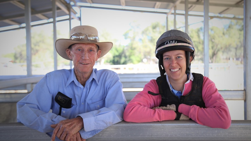 An older man and a younger woman lean on a stable fence wearing racing gear