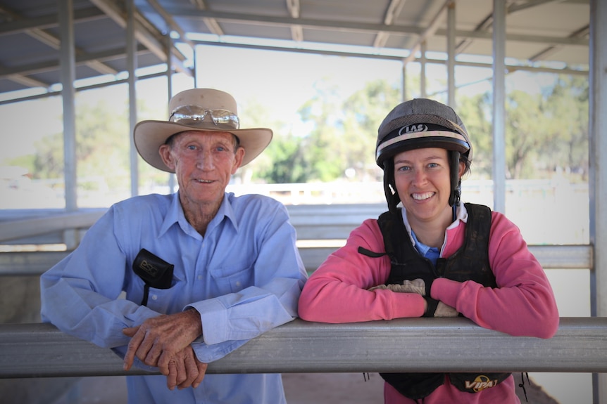 An older man and a younger woman lean on a stable fence wearing racing gear