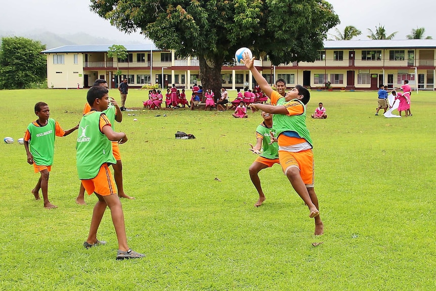 A group of young boys playing touch rugby with fellow students with the school in the background.