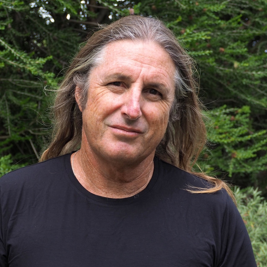 A man with long hair standing in front of green foilage smiling