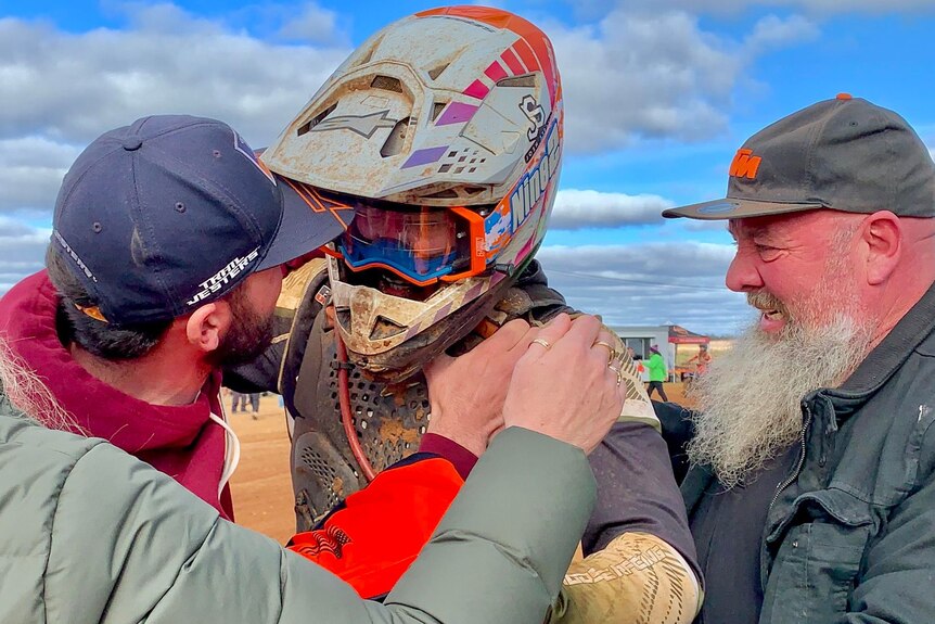 two emotional men hug a dirt bike rider with his helmet on