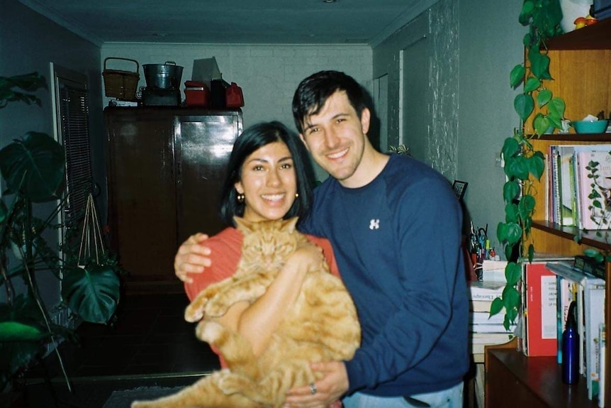 A man and woman stand hug holding a ginger cat, laughing.