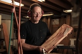 Bob Jolliffe smiles at the camera while holding a plank of timber, wearing ear muffs and a black shirt.