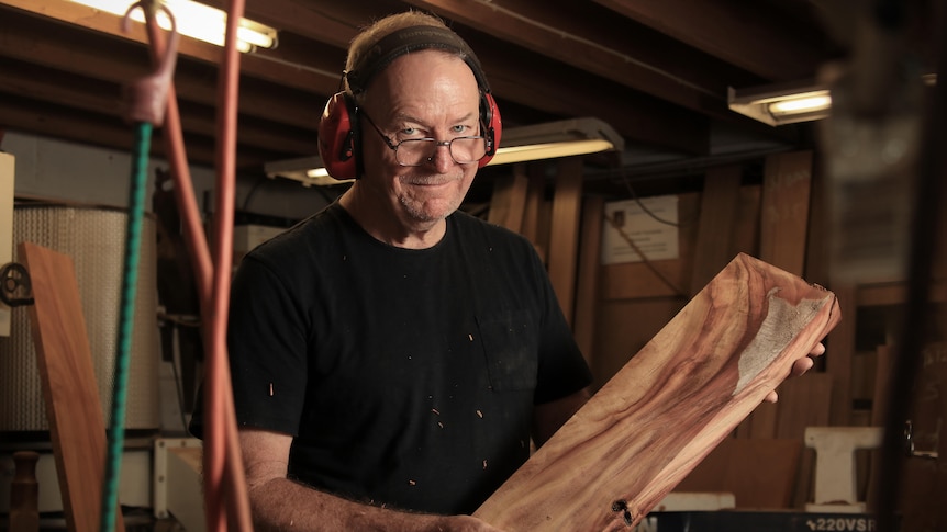 Bob Jolliffe smiles at the camera while holding a plank of timber, wearing ear muffs and a black shirt.