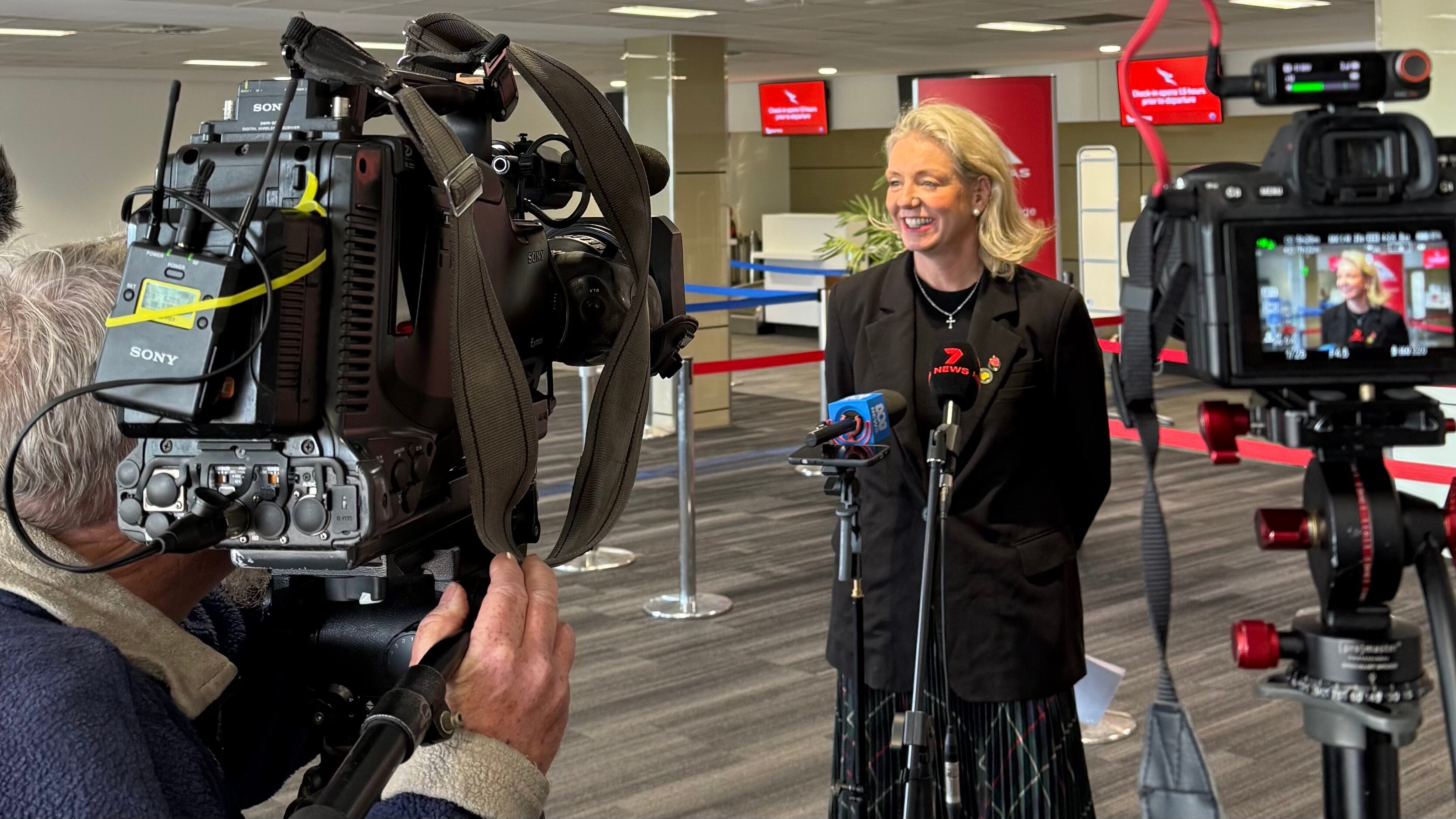 Uma mulher diante de várias câmeras com um terminal de aeroporto atrás dela. 
