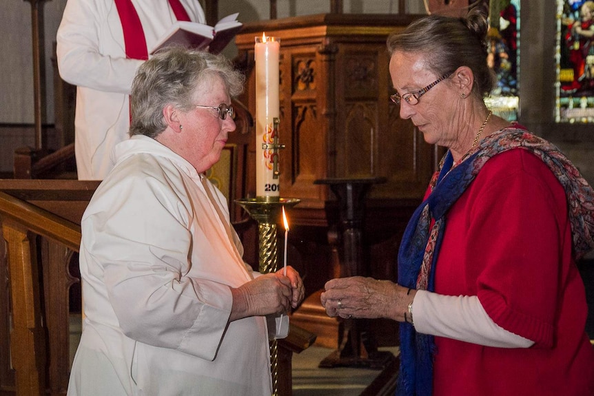 A woman in white church robes holds a lighted taper while another woman holds a candle (hidden in her hands) ready to be lit