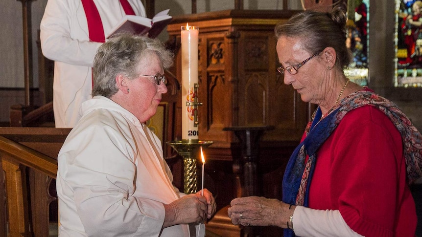 A woman in white church robes holds a lighted taper while another woman holds a candle (hidden in her hands) ready to be lit