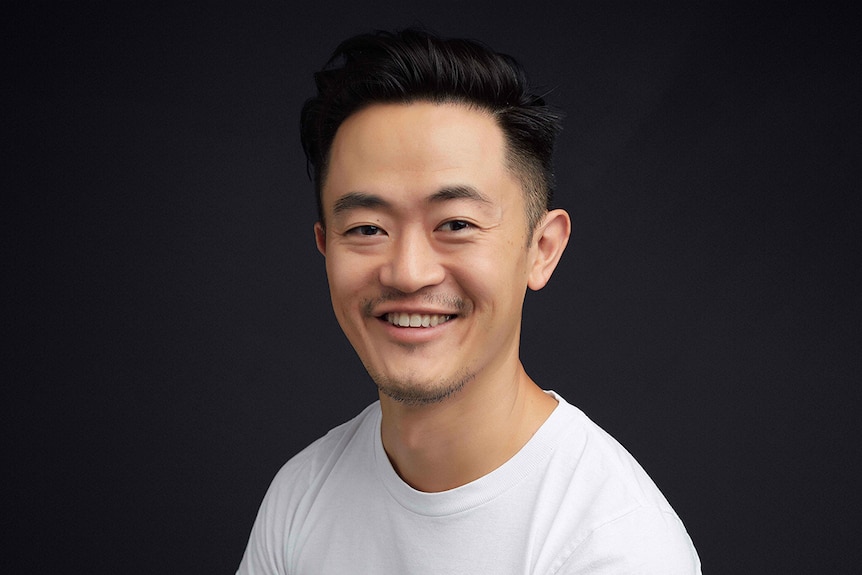 A man wearing white t-shirt and smiling towards camera is seated in front of dark grey wall.