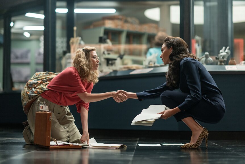 Kristen Wiig and Gal Gadot in 80s office clothes, crouching on office floor over dropped papers, shaking hands.