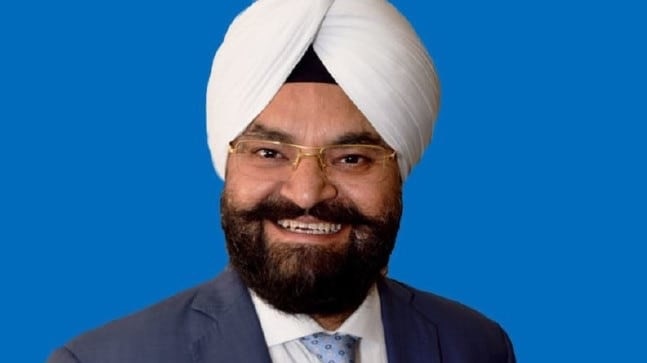 Gurpal Singh smiles for a portrait photograph, in front of a blue background.