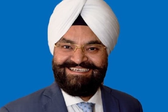 Gurpal Singh smiles for a portrait photograph, in front of a blue background.