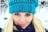 A photo of a young blonde-haired woman wearing a blue beanie in a snowy location.
