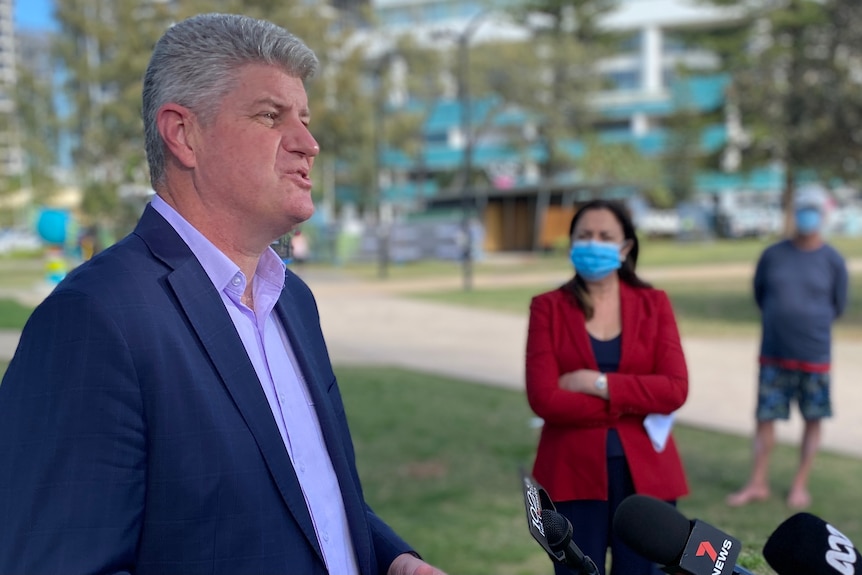 A man in a dark suit stands in a park speaking to the media.