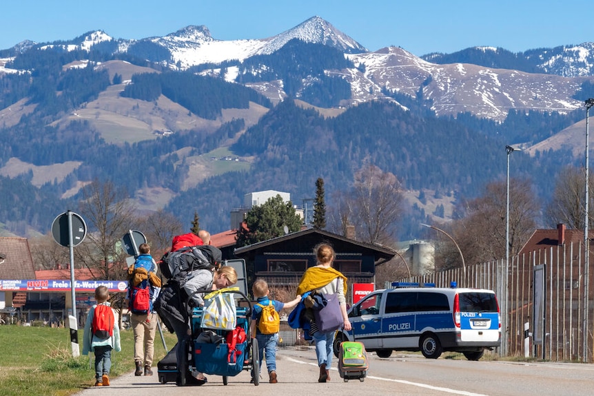 Against an alpine backdrop, you see two young families walk across a border crossing.