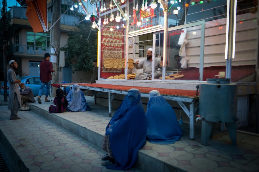 Women in blue burqa sit on the concrete steps outside a bakery stall. 