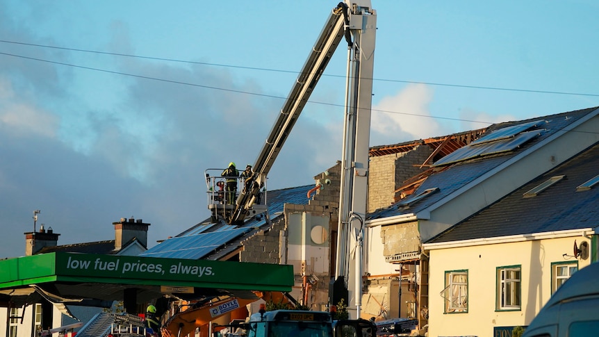A person on a cherry picker is lifted above the destroyed roof of a petrol station. 