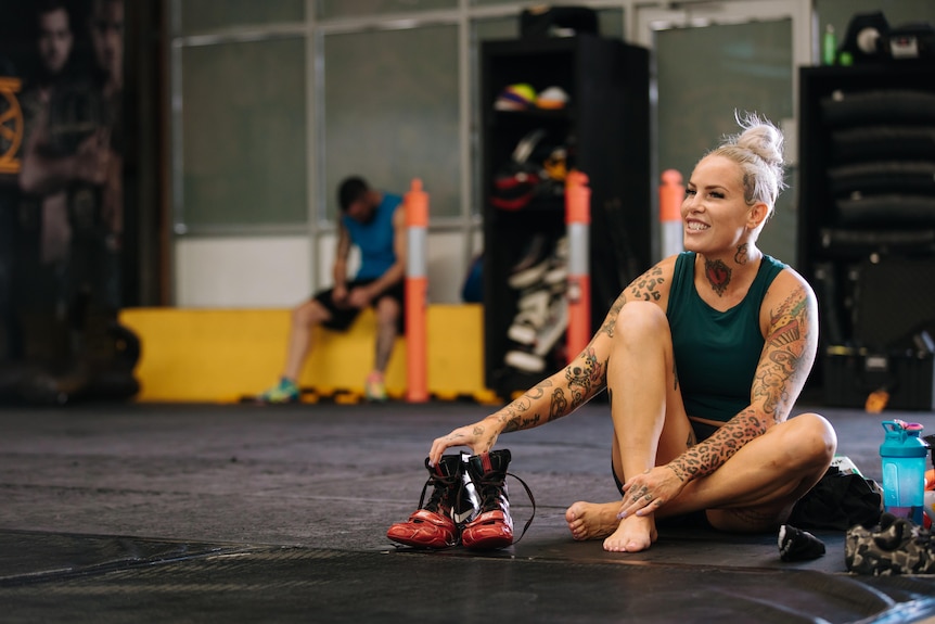 A woman sits on the floor of a gym, holding onto her shoes, smiling off into the distance.