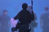 An officer in the foreground walks towards other authorities with his weapon raised.