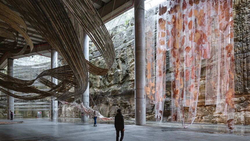 A structure of made of bamboo in the shape of a wave form and coloured fabrics each hang from the ceiling of a cavernous space