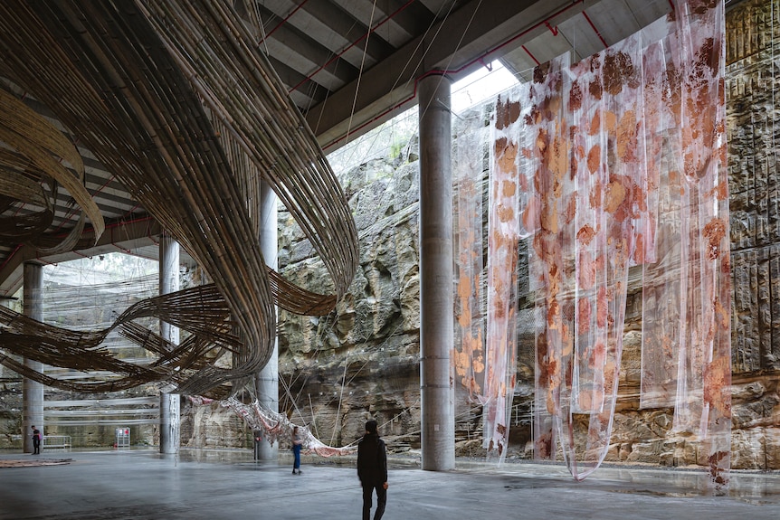 A wave-like bamboo structure and colorful fabrics each hang from the ceiling of a cavernous space