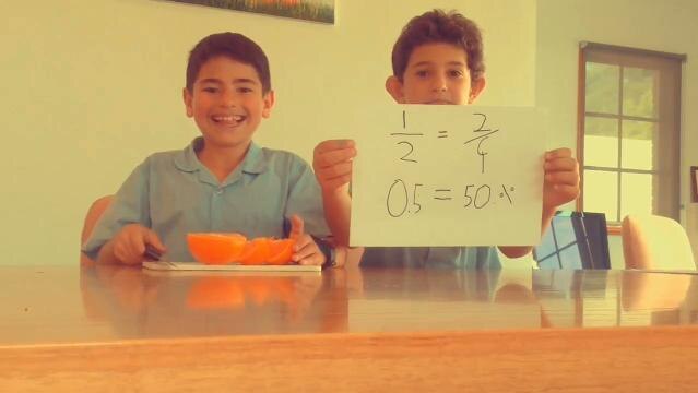 Two boys sit at table, one has cut oranges, the other holds up paper with written sums