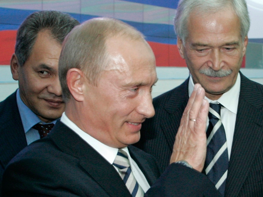 Vladimir Putin waves while two men stand behind him, glancing sideways. One has a grey brush-like moustache
