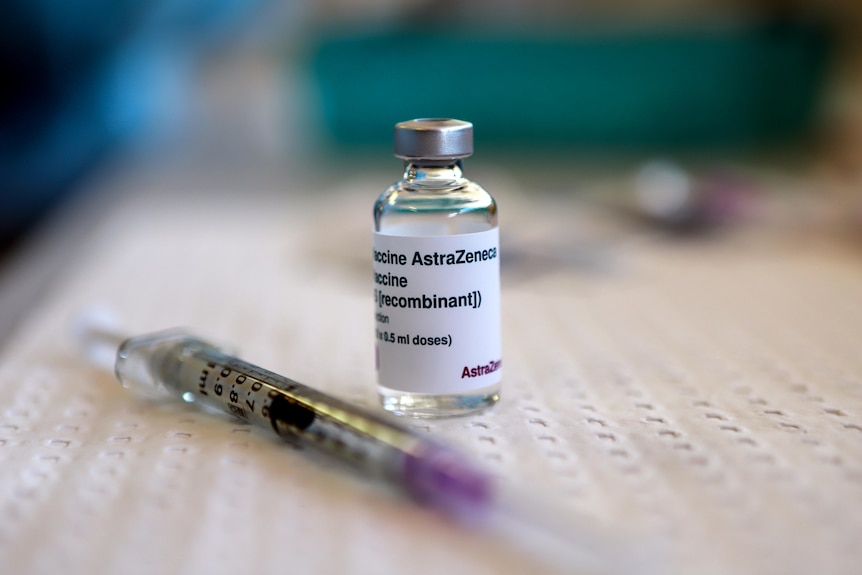 Small glass vial with label reading Astra Zeneca sits on bench next to a syringe
