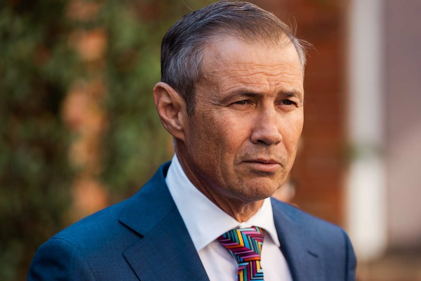 A headshot of Roger Cook standing in a brick courtyard wearing a suit and multicoloured tie.