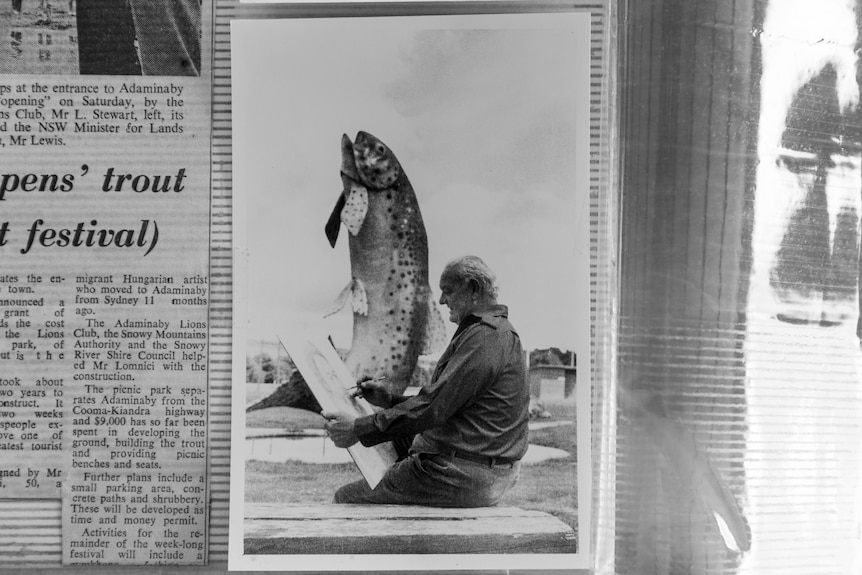 A black and white image of an artist sitting beside a large statue of a big rainbow trout