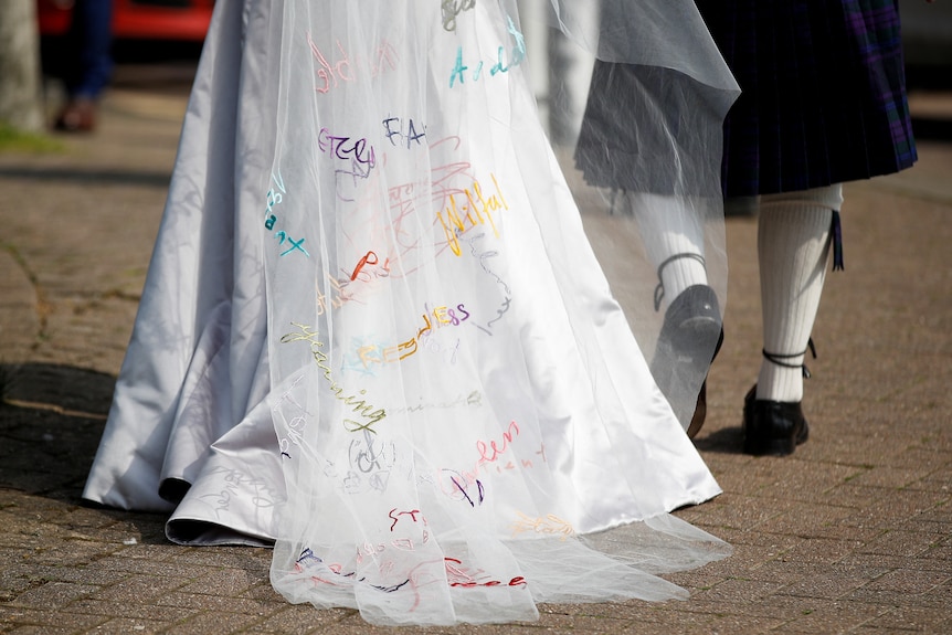 A close up on a white veil that reaches the floor. Colourful words are scrawled across it like 'brave' and 'wlilful'