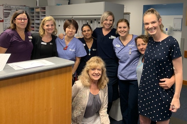 A blonde woman in a dark dress with a big smile, surrounded by hospital staff.