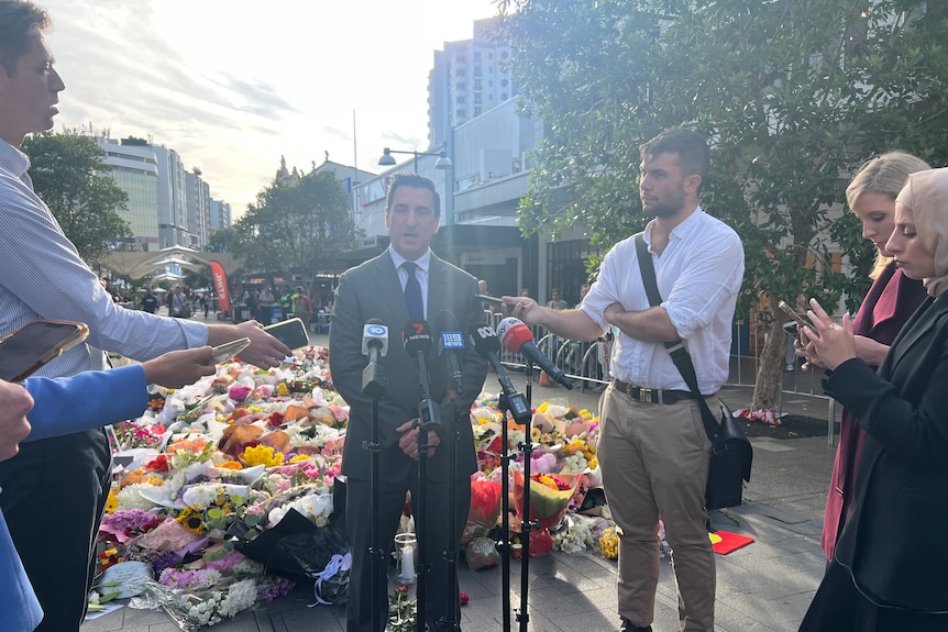 Elliott Rusanow stands with his back to the makeshift memorial in front of a row of microphones