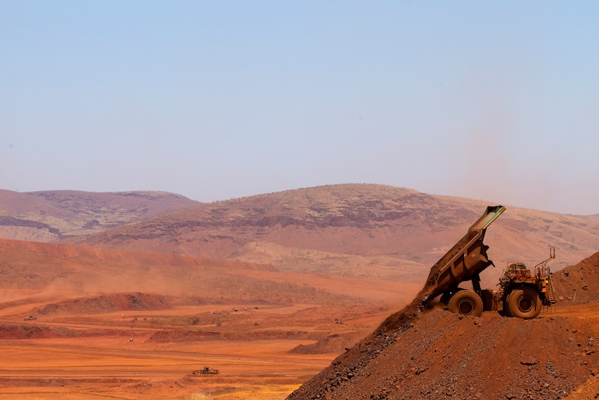 A dump truck emptying a pile of red dirt into a very large mine - there are hills in the background.