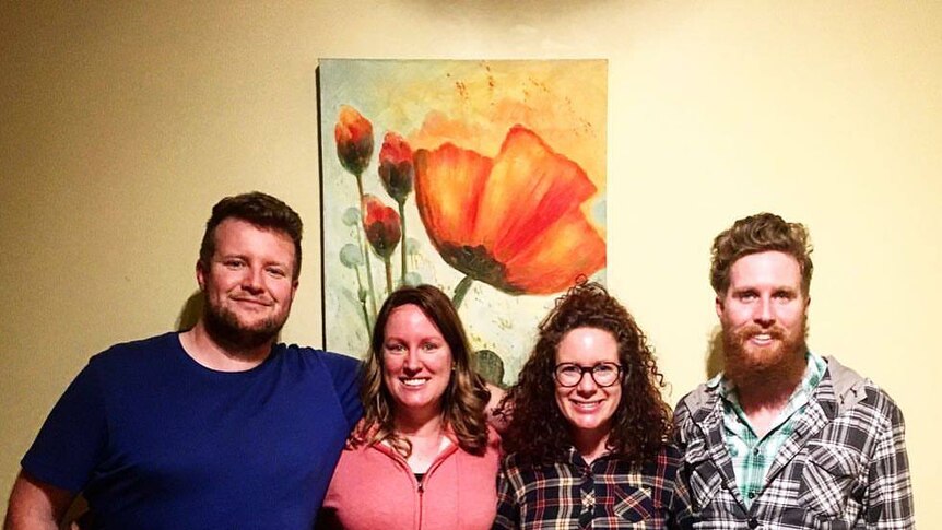 Four people stand together, smiling at the camera. An artwork of a flower sits on the wall behind them.