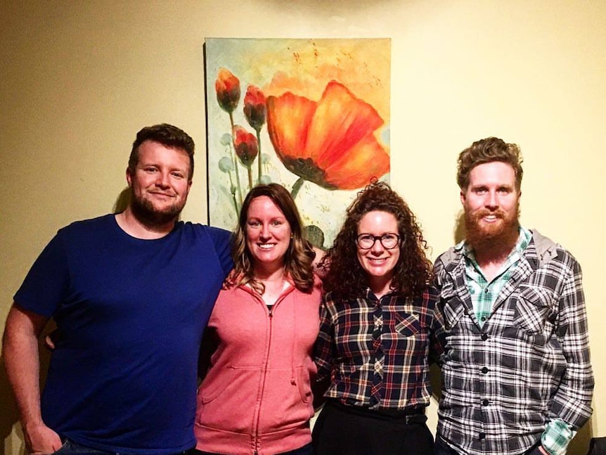 Four people stand together, smiling at the camera. An artwork of a flower sits on the wall behind them.