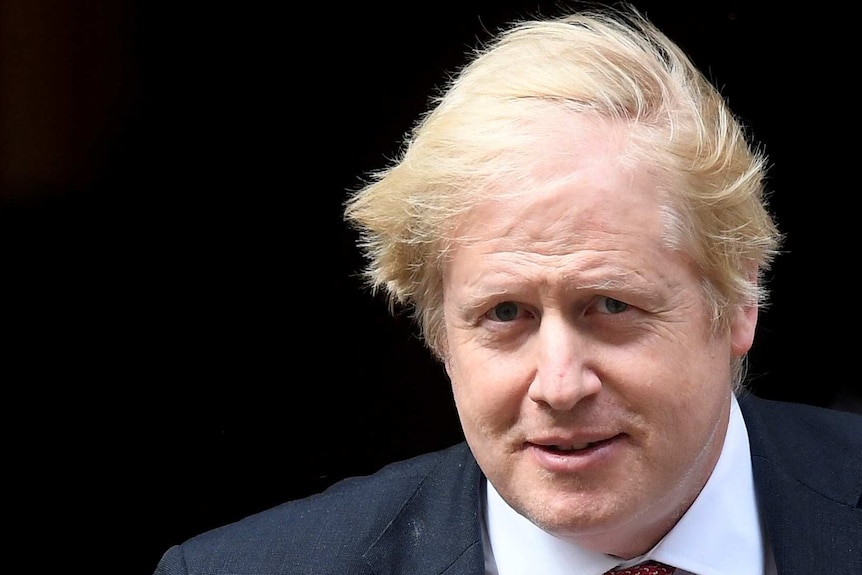 A close-up of a wind-swept man with fair hair, wearing a dark suit, white shirt and red tie.